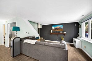 Ein modernes Wohnzimmer mit einem grauen Ecksofa, einem kleinen Tisch, einer Lampe, einem Fernseher auf einem Holzregal und einem Fenster mit Blick.