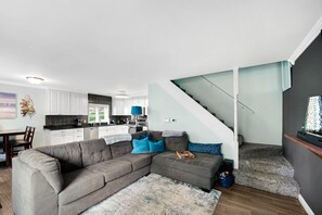 Ein modernes Wohnzimmer mit Ecksofa, einer Treppe und einer Küche im Hintergrund.