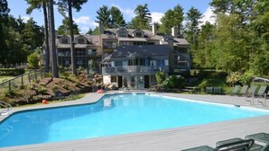 A swimming pool with lounge chairs, a wooden deck, and a large building with multiple balconies in the background. - Samoset Lakeview Gem - Vibrant Condo Escape! (Gilford)