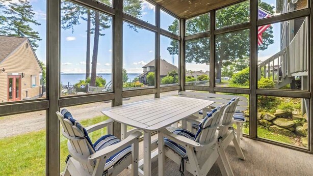 A covered patio with a table and chairs, large windows offering a view of the sea and surrounding houses. - Samoset Lakeview Gem - Vibrant Condo Escape! (Gilford)