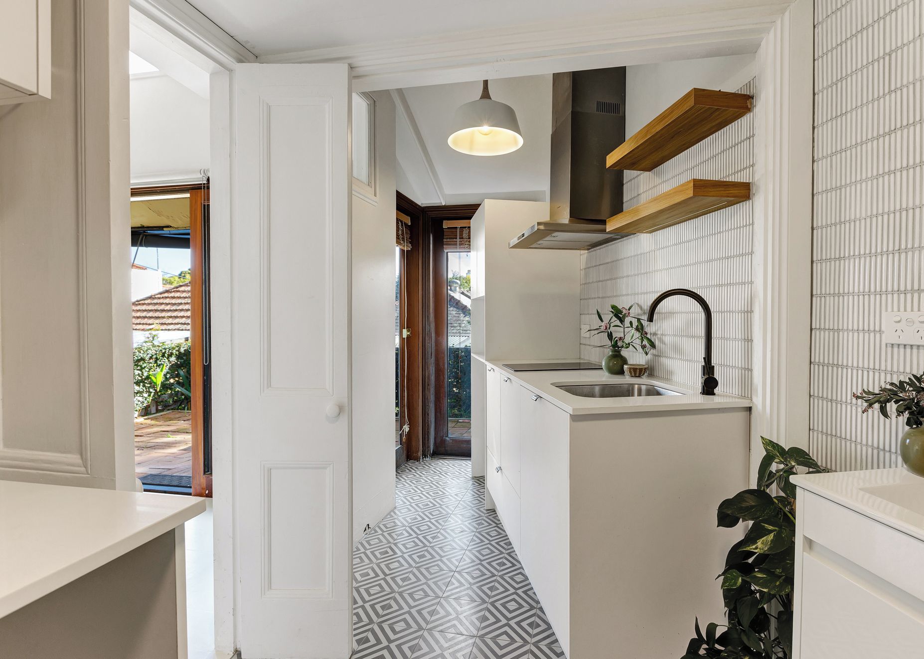 A narrow hallway with a patterned floor, a kitchen area with a sink and a hanging light fixture, and a view into another room.