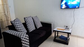 Musta sohva, jossa on kuviollisia koristetyynyjä, pieni pöytä, jonka päällä on lehti, ja seinälle asennettu televisio.