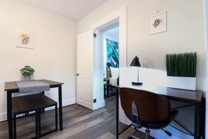A modern, well-lit room with a dark wood desk, a chair, and a small table with a plant. There is a door leading to another room with a colorful painting. - Wynwood Apartments (Miami)