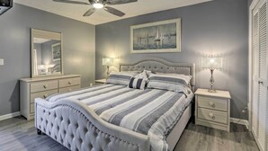 Une chambre à coucher avec une tête de lit capitonnée, un grand lit avec une literie à rayures, deux tables de chevet avec des lampes, un ventilateur de plafond et un tableau encadré accroché au mur.