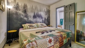 A bedroom with a large bed, a forest mural on the wall, a small round table, and a door with a mirror. - King Suite & Private Hot Tub Great Location (Gatlinburg)