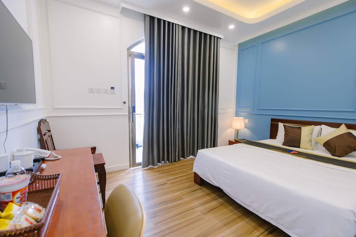 A hotel room with a wooden floor, a bed with white bedding, a desk with a telephone and basket, and a window with curtains.