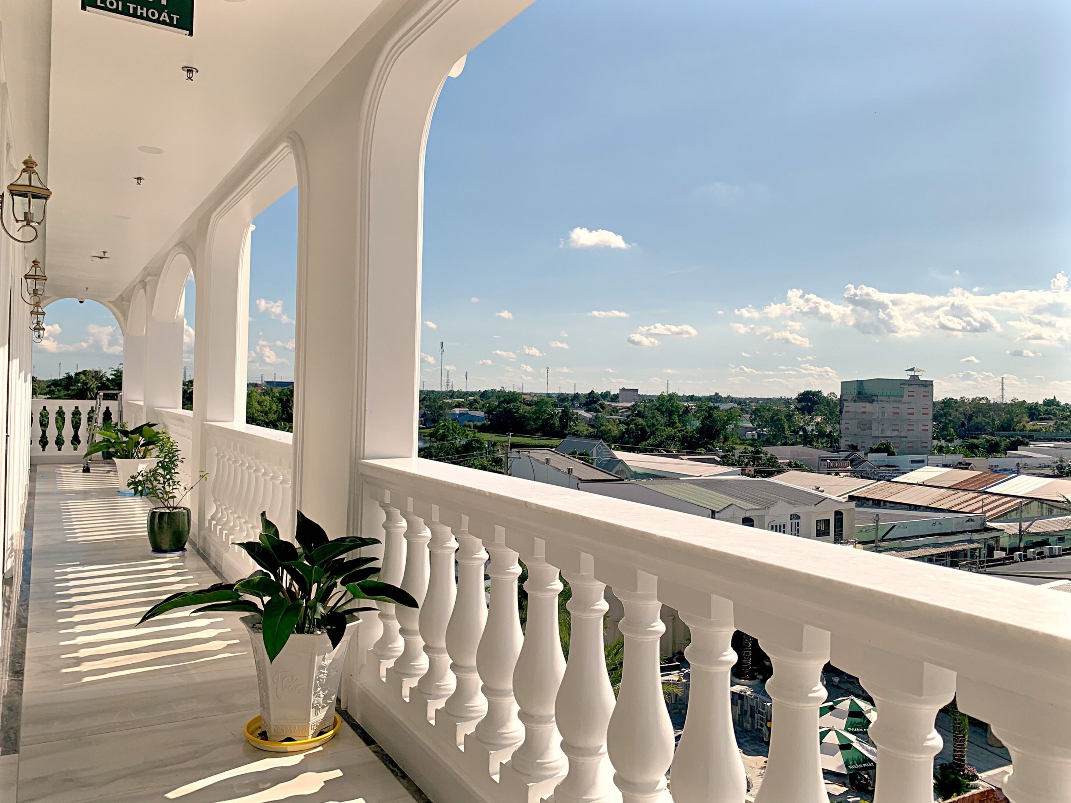 A balcony with white balustrades and potted plants, overlooking a cityscape with buildings and greenery.