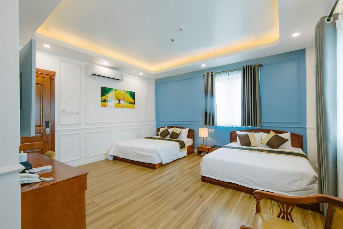 A hotel room with two beds, a wooden desk, a telephone, and a window with curtains.