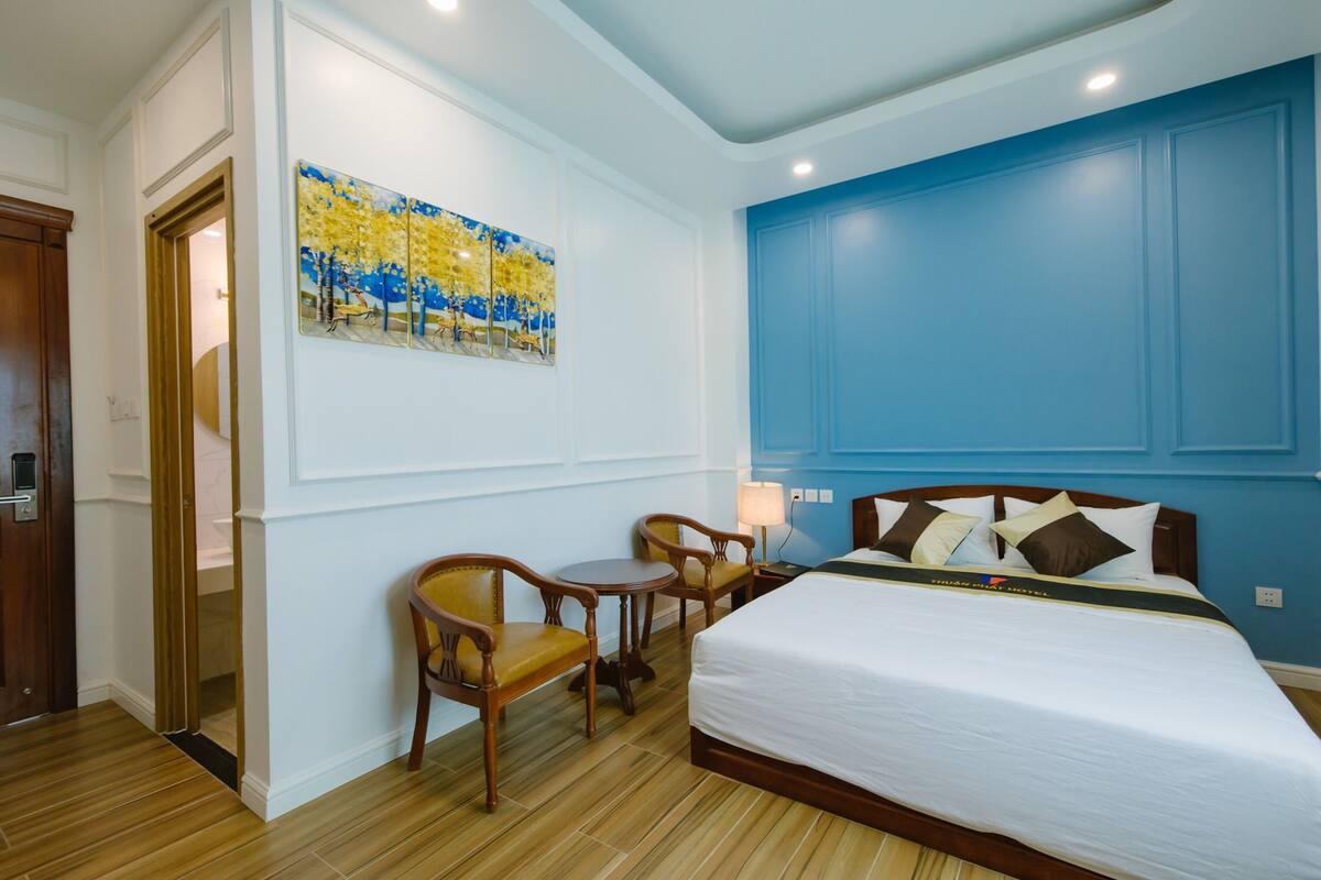 A hotel room with a large bed, two wooden chairs, a small table, a painting on the wall, and a blue accent wall.