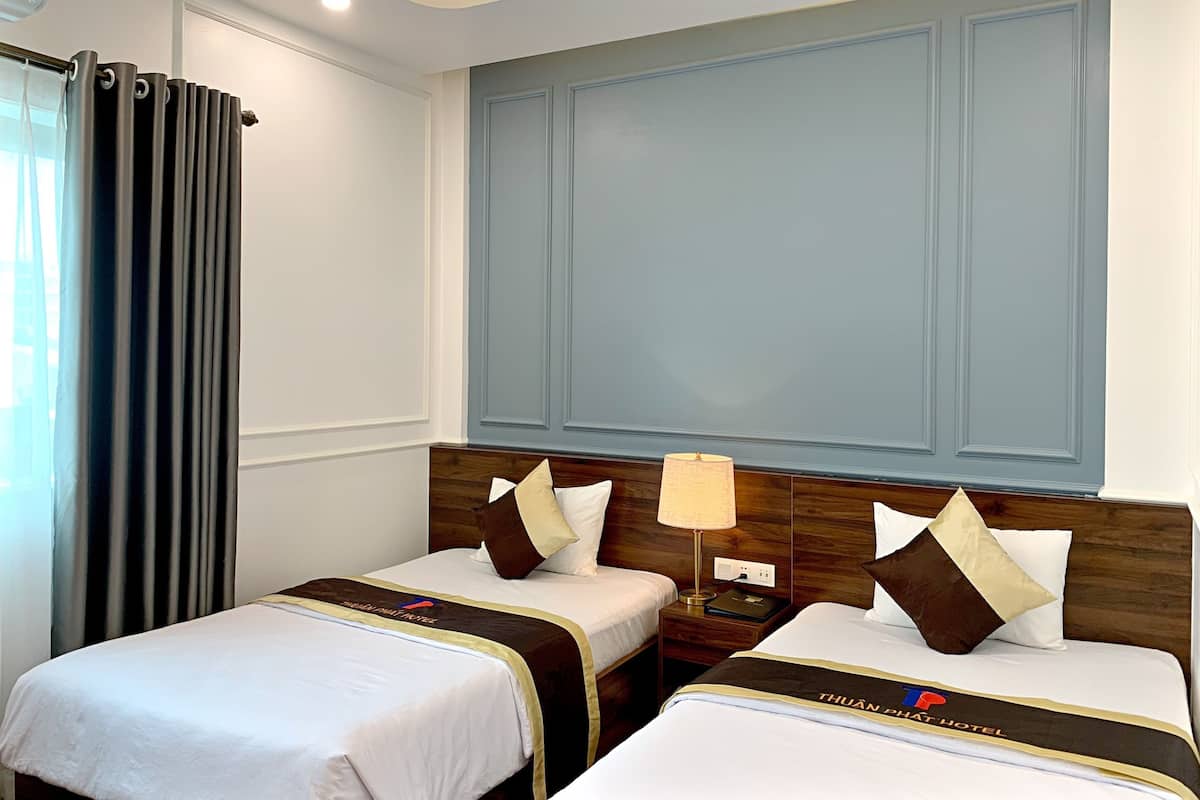 A hotel room with two beds, each with a headboard, a bedside table with a lamp, and a curtain on the window.