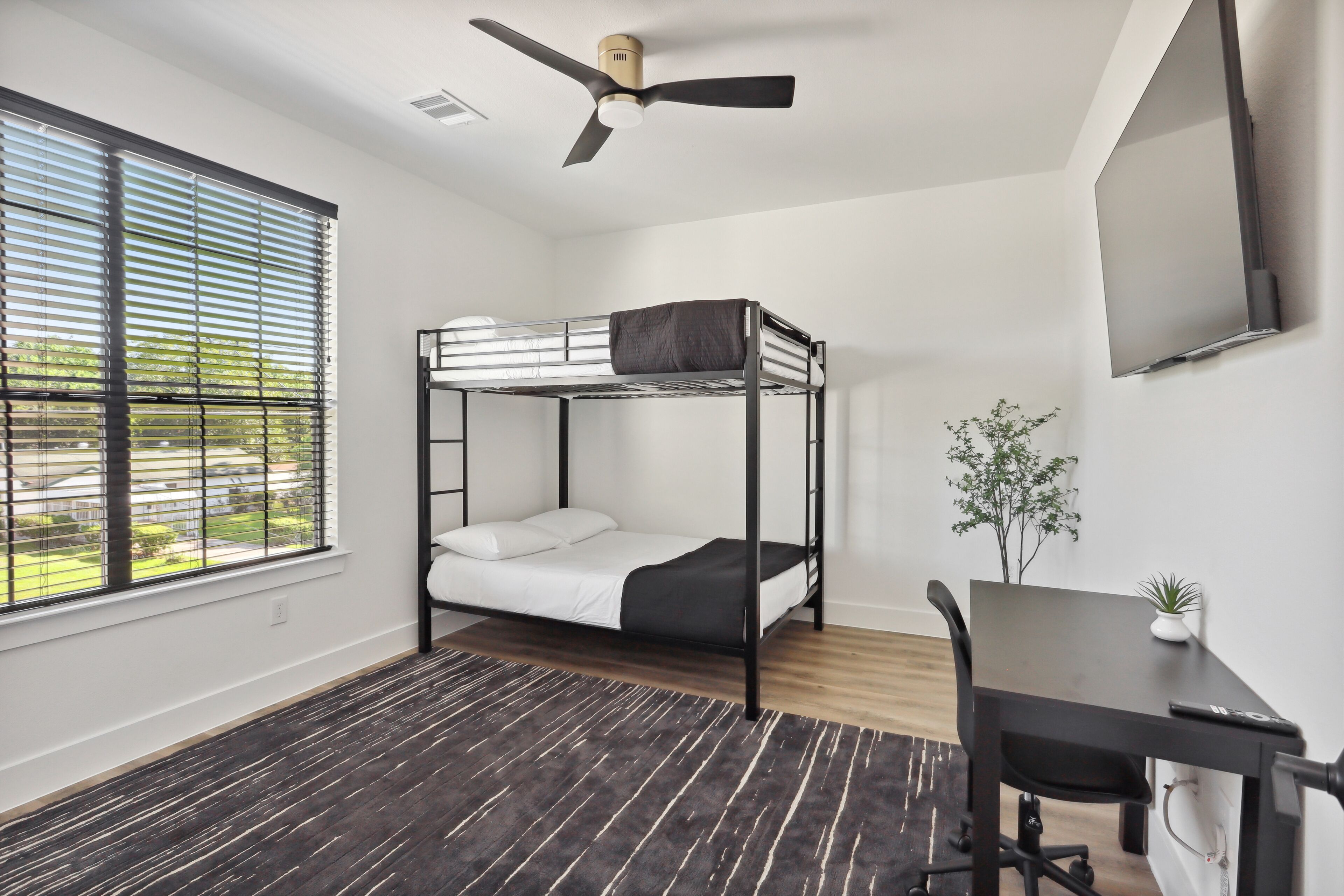 A room with a bunk bed, a desk with a chair, a ceiling fan, a window with blinds, and a small plant.