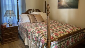 Ein ordentlich bezogenes Bett mit floralem Überwurf, ein Nachttisch aus Holz mit einer Lampe und ein Gemälde an der Wand.