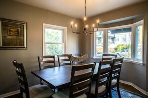 Uma sala de jantar com uma mesa de madeira escura e seis cadeiras, um candelabro e janelas amplas com vista para o exterior.