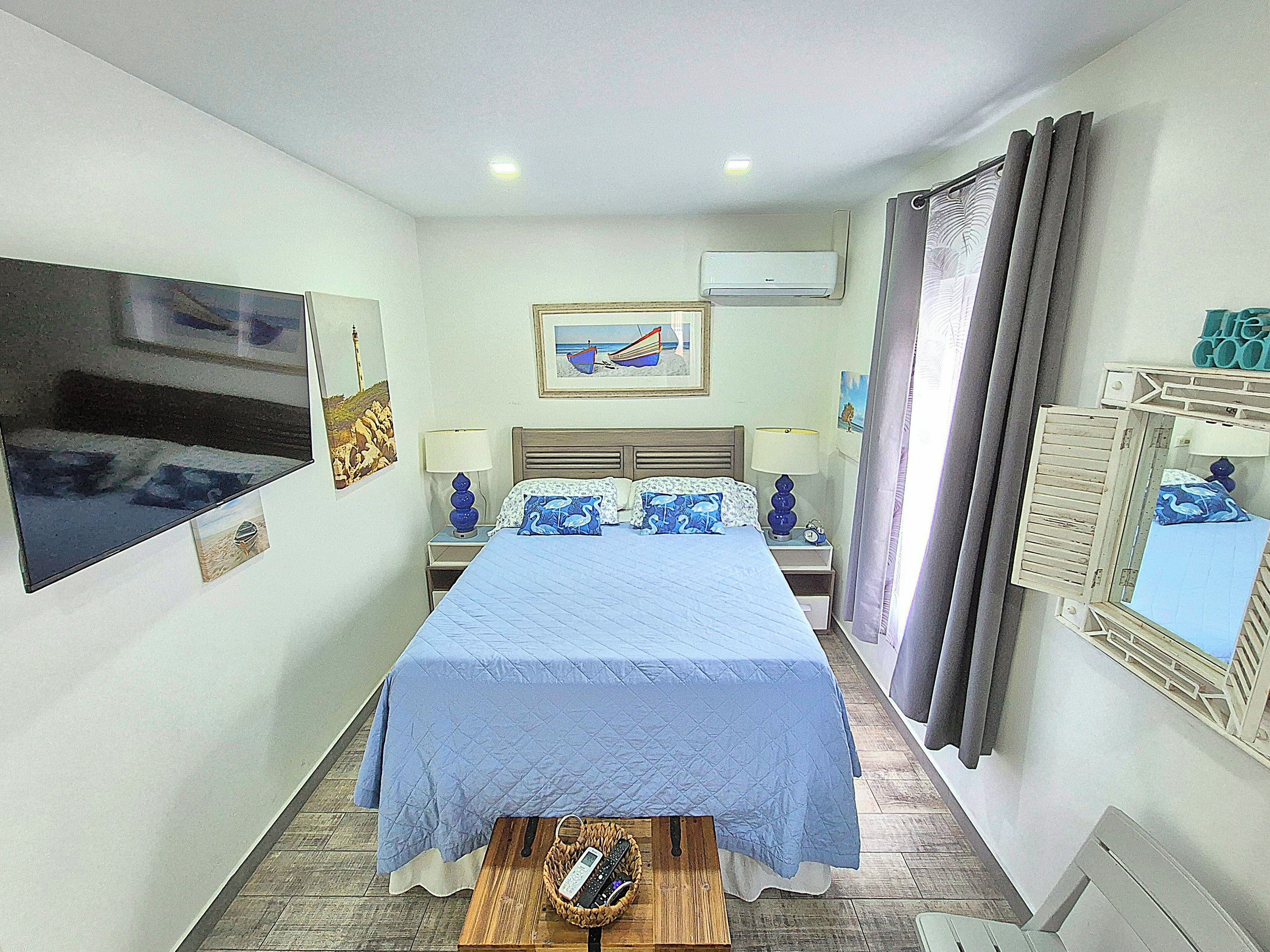 A hotel room with a bed, bedside tables, a TV, artwork, and an air conditioning unit.