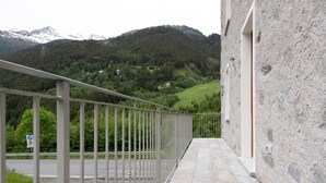 A balcony with a view of a mountainous landscape, a paved walkway, and a building with a stone facade.