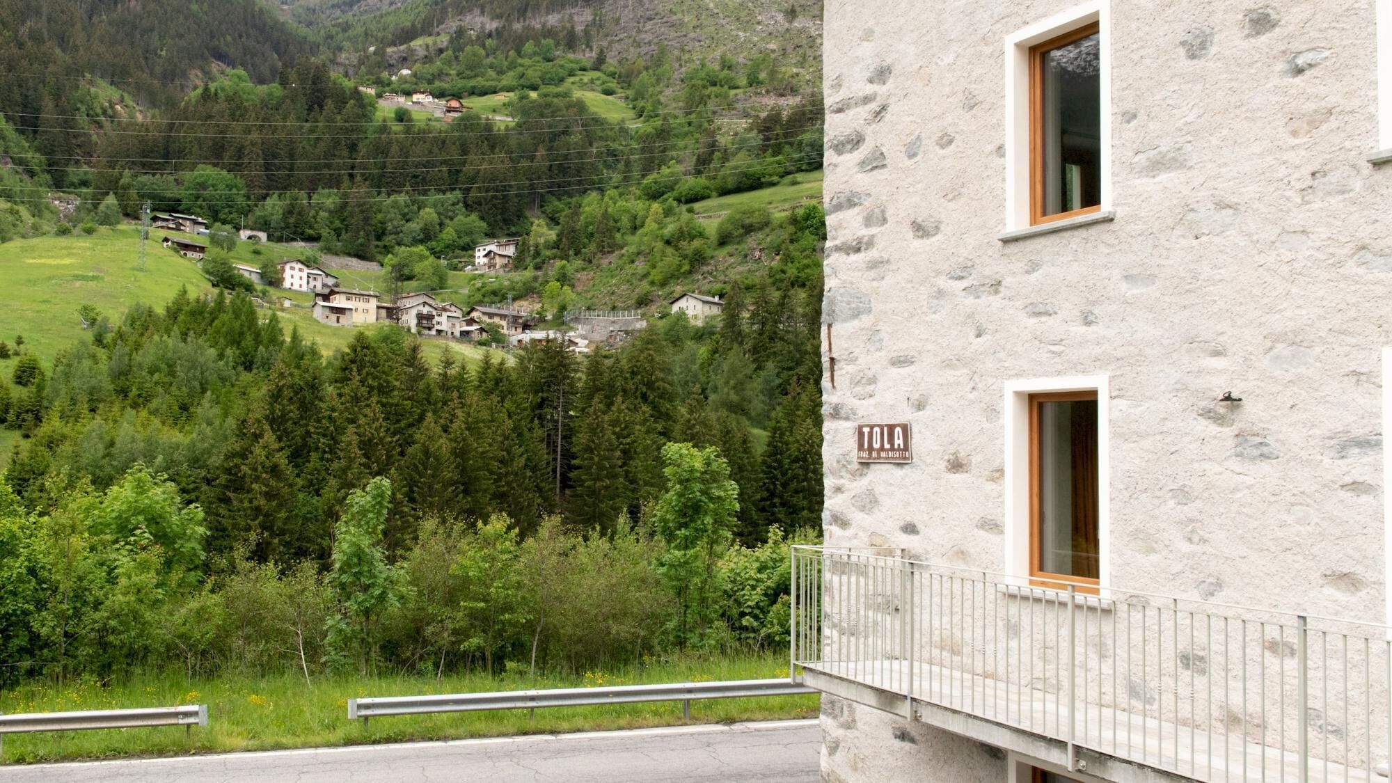 A stone building with a balcony, a sign reading "TOLA," and a view of a green valley with houses.