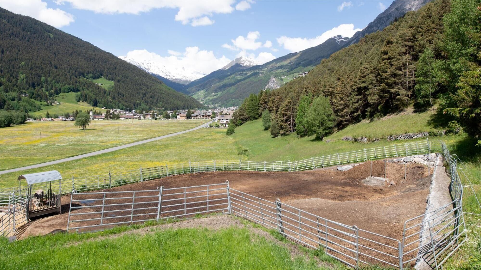 A fenced outdoor area with a dirt ground, surrounded by green fields and mountains in the background.