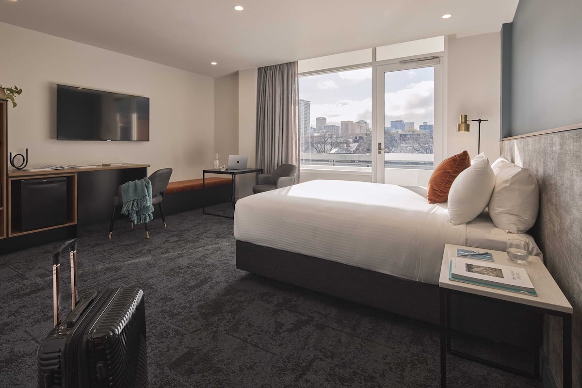 A modern hotel room with a large bed, a desk with a laptop, a TV, and a view of the city.