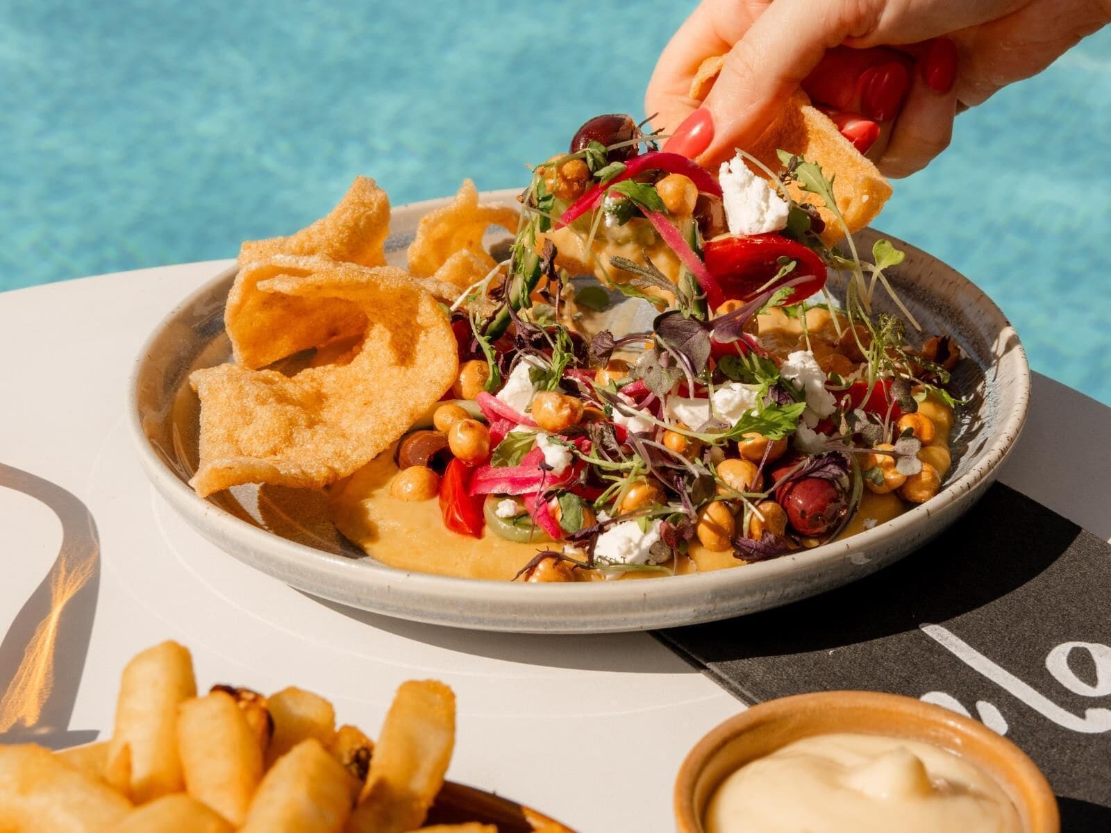 Un tazón de comida con diferentes ingredientes y totopos, una mano sumergiendo un totopo en salsa, y una piscina al fondo.