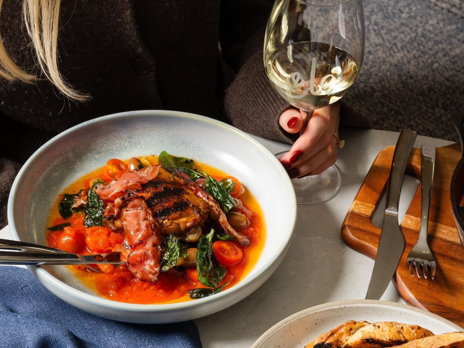 A bowl of soup with vegetables and meat, a wine glass, and cutlery on a table.
