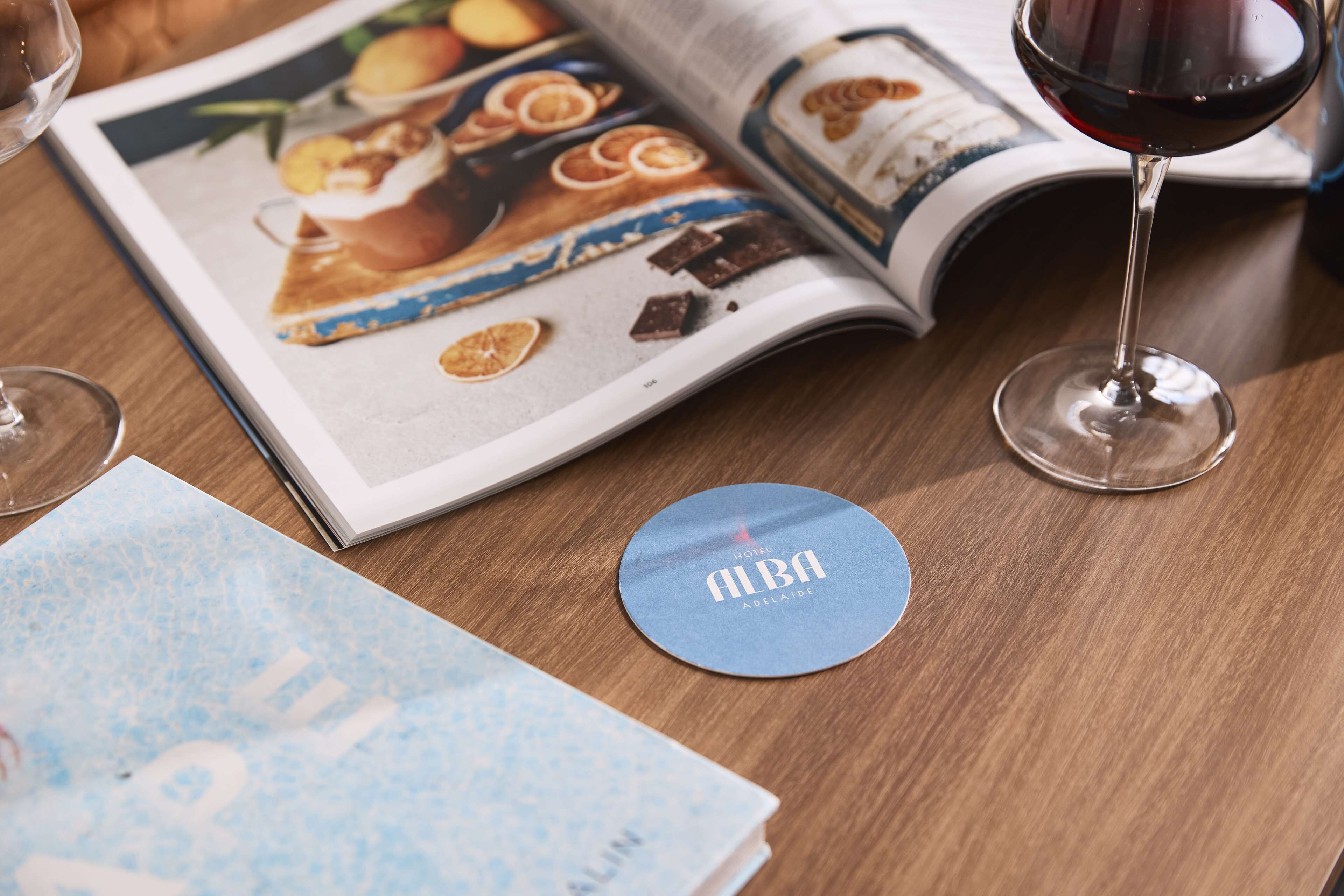 A magazine with a round blue coaster labeled "ALBA HOTEL ADELAIDE" on a wooden table.