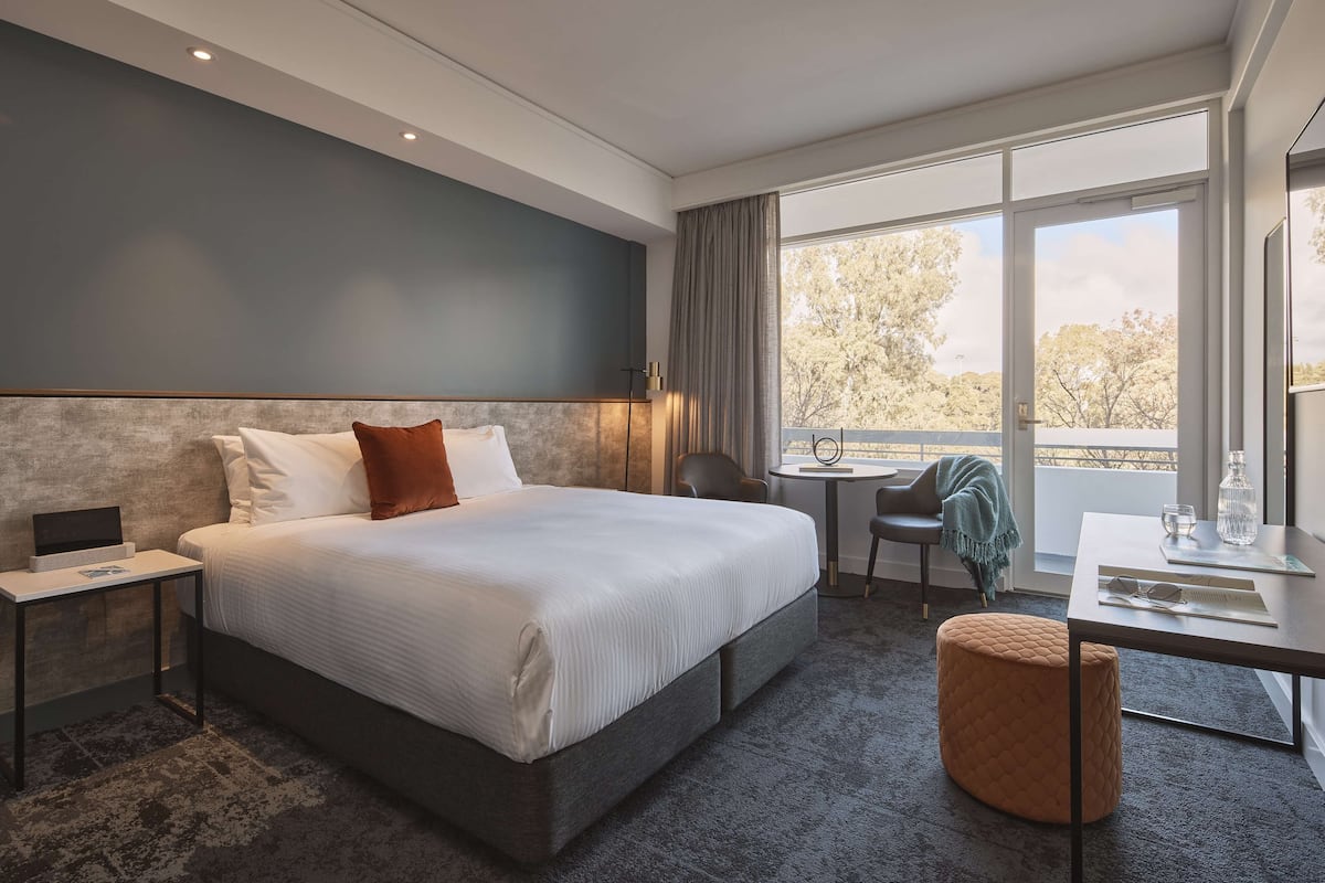 A modern hotel room with a large bed, bedside table, and a small desk with a chair.
