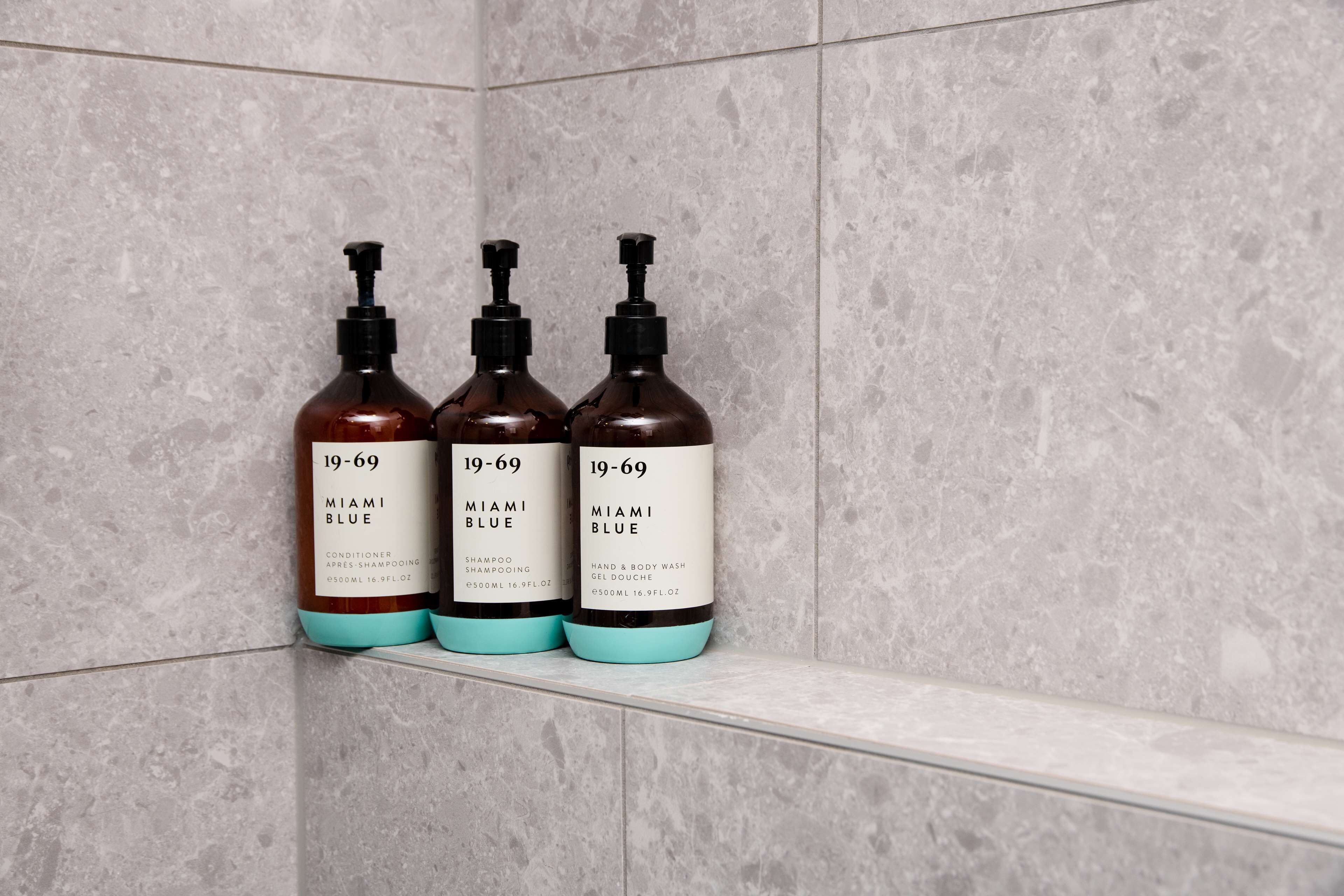 Three bottles of Miami Blue shampoo and conditioner on a shelf against a tiled wall.