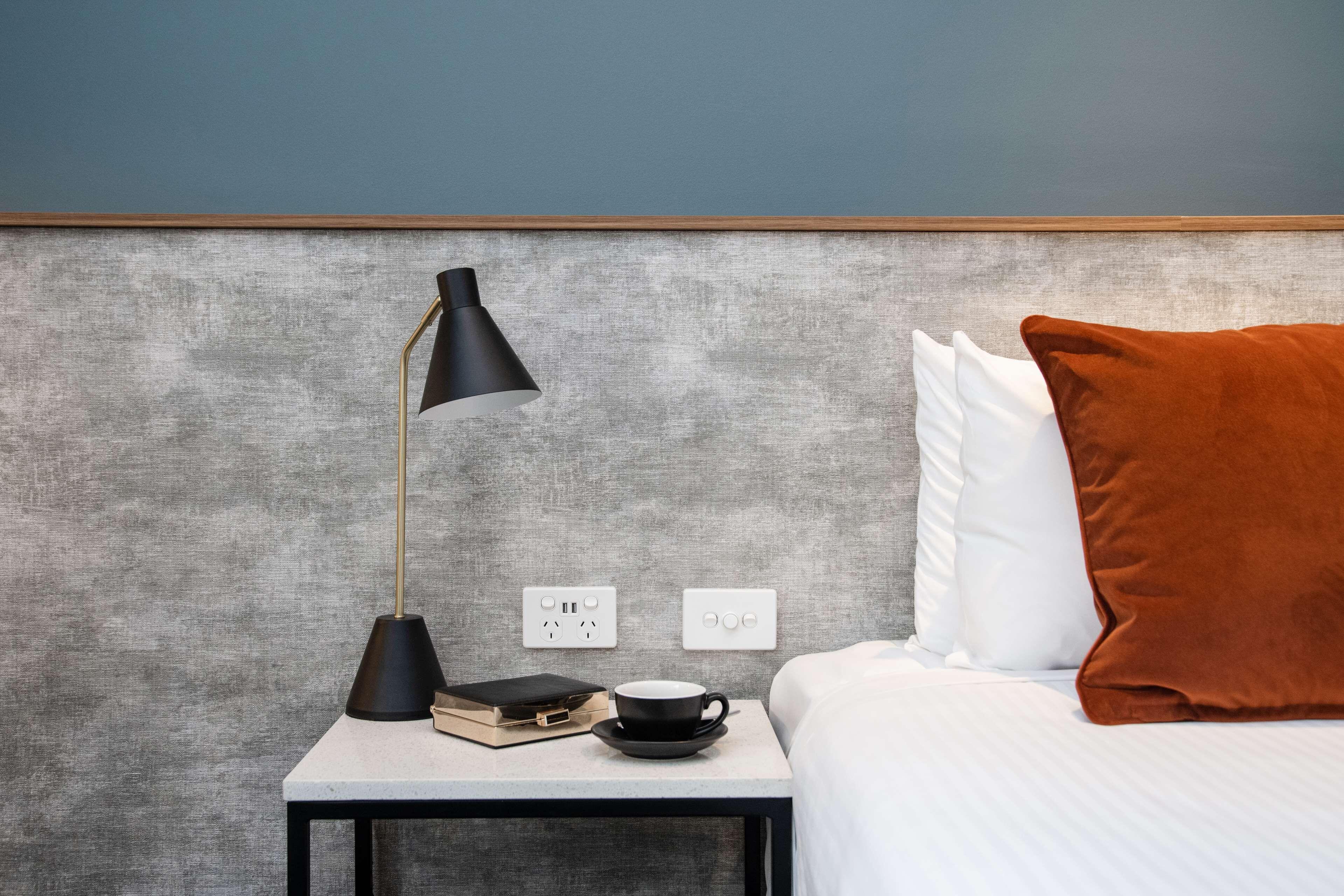 Quarto de hotel bem organizado, com uma cama, mesa de cabeceira com abajur, uma xícara e um livro.