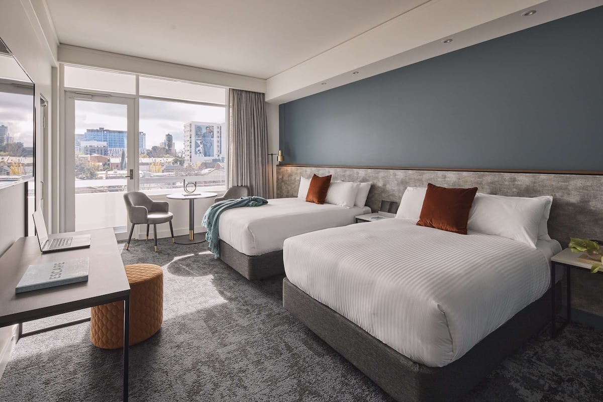 A modern hotel room with two beds, a desk with a laptop, a chair, and a view of the city.