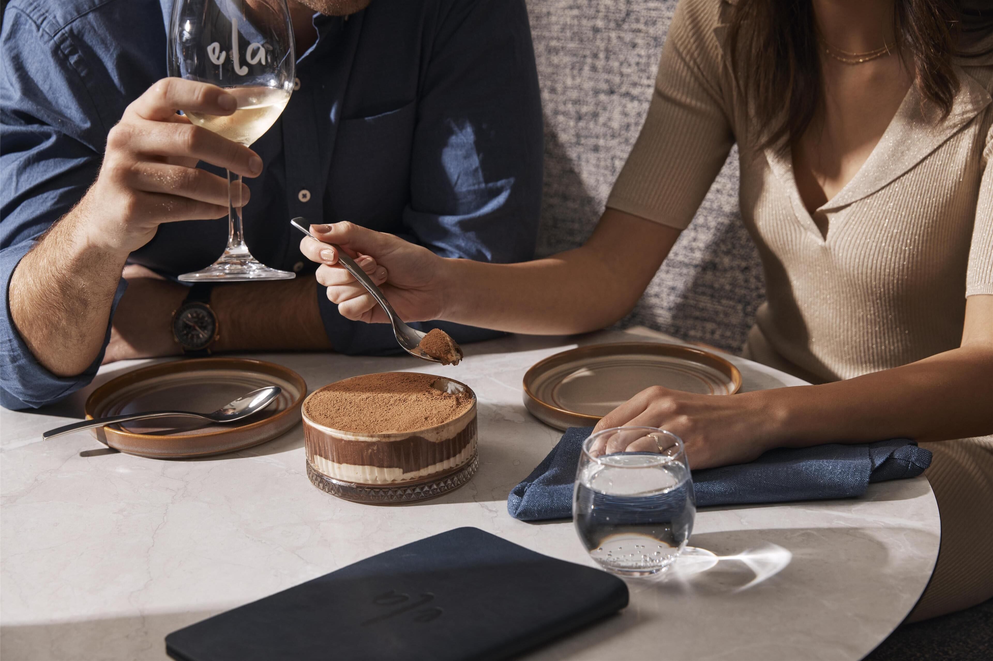 Una mesa de comedor con un postre, una copa de vino blanco y un teléfono inteligente.