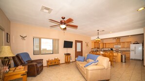 A spacious living room with a beige sofa, a brown leather chair, and a wooden coffee table. There is a kitchen area with wooden cabinets and a refrigerator. - Foothills Home (Yuma)