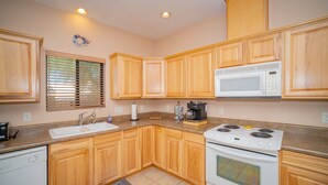 A kitchen with wooden cabinets, a white microwave, a stove, a sink, and a window with blinds. - Foothills Home (Yuma)
