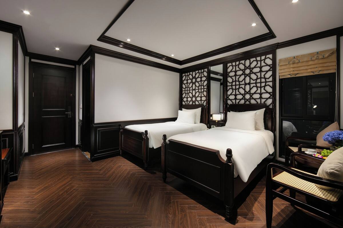 Une chambre d’hôtel avec deux lits, un plancher en bois, une armoire de couleur sombre, un paravent décoratif et une fenêtre avec vue.