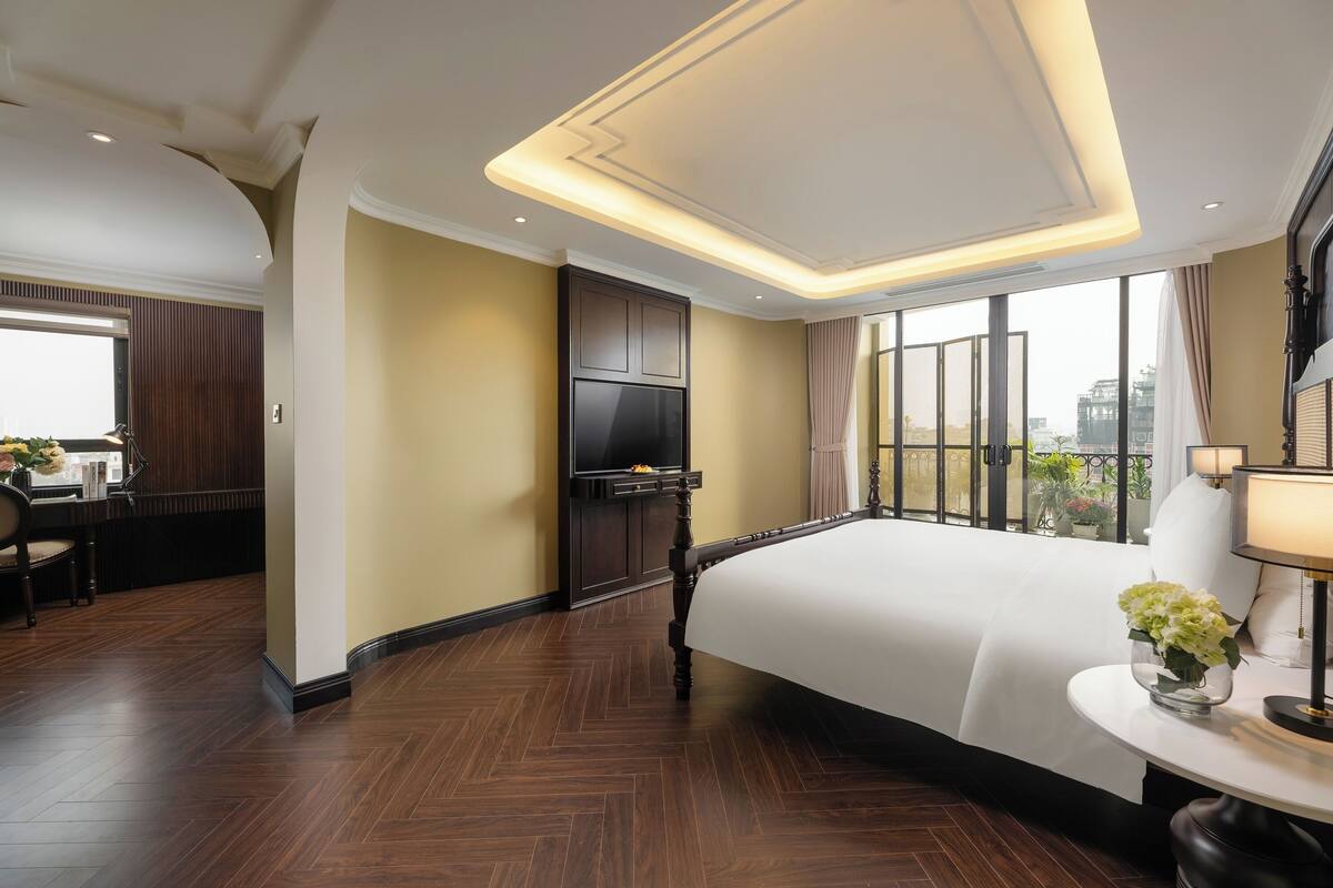 Une chambre d’hôtel dotée d’un grand lit, d’un téléviseur fixé au mur, d’un bureau avec une chaise et offrant une vue sur la ville grâce à de grandes fenêtres.