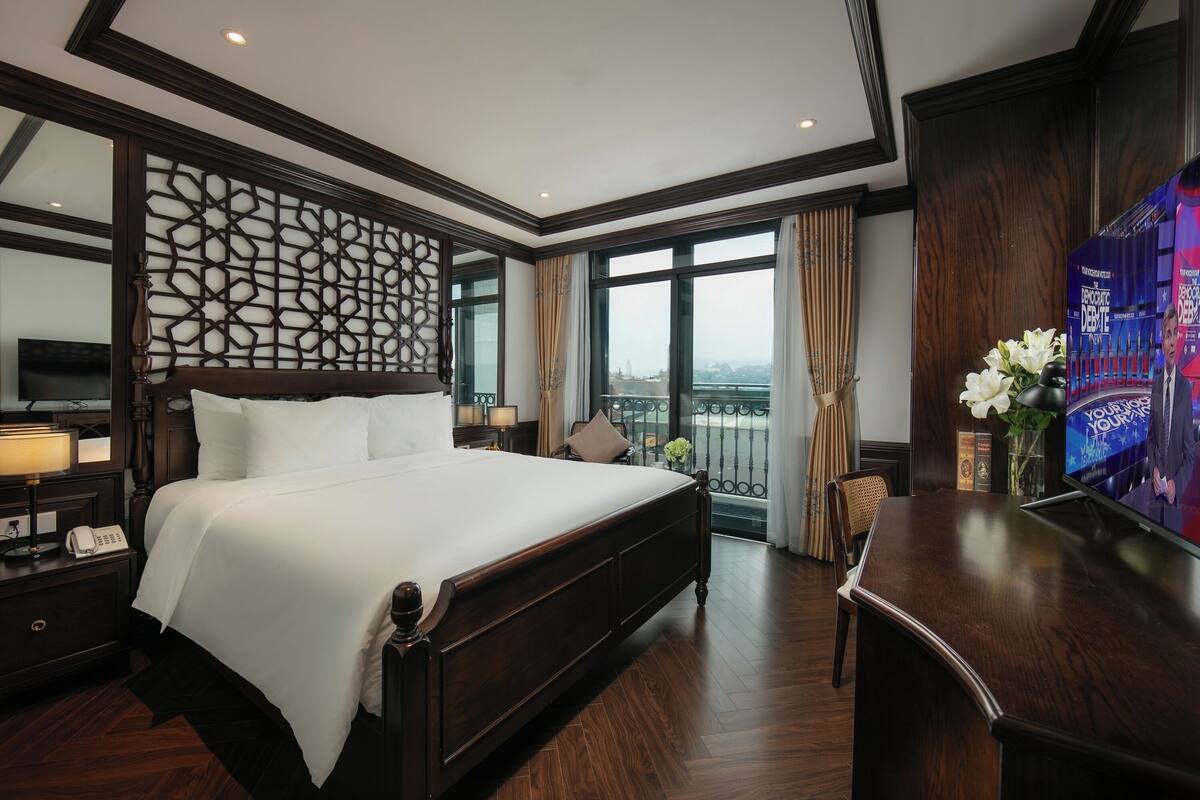 Une chambre d’hôtel avec un grand lit, une tête de lit en bois, un bureau, une télévision et une vue sur la ville grâce à de grandes fenêtres.