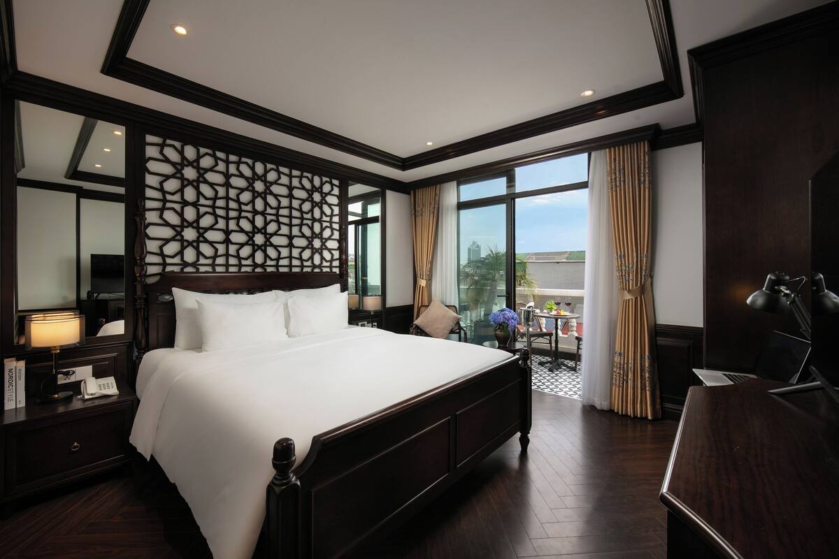 Une chambre d’hôtel spacieuse, dotée d’un grand lit, d’un bureau, d’une chaise, d’une lampe, d’un miroir, d’un panneau décoratif mural et offrant une vue sur la ville grâce à de grandes fenêtres.