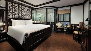 Una habitación de hotel moderna con una cama grande, un box de ducha con paredes de vidrio, una zona de estar pequeña y un escritorio con lámpara.