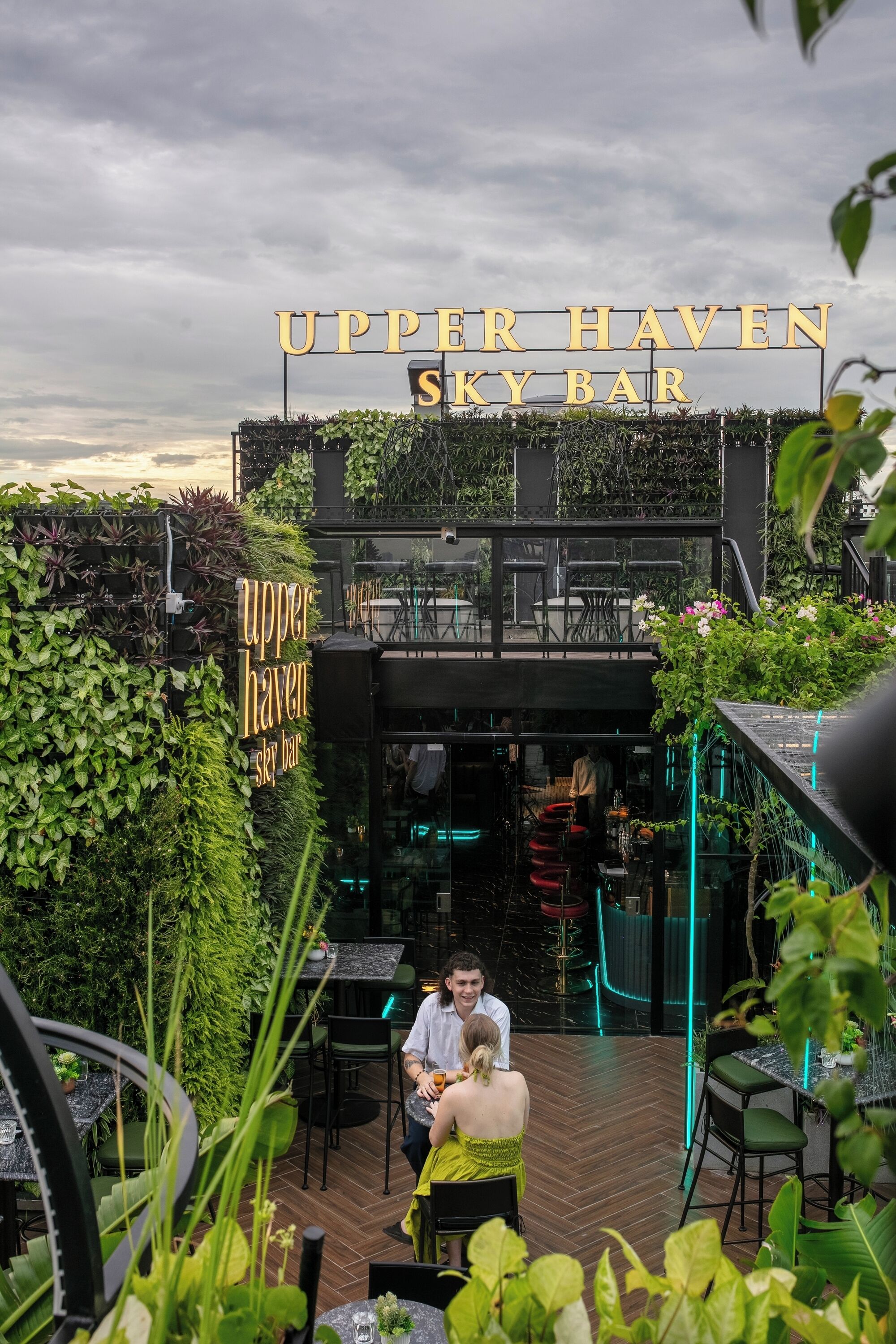 Un espace extérieur aménagé avec des tables et des chaises, entouré d’une végétation luxuriante et agrémenté d’une enseigne indiquant « Upper Haven Sky Bar ».
