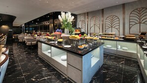 Un vestíbulo de hotel moderno con piso de mármol, un bufé central con diversos alimentos y un área de descanso con sofás y sillones.