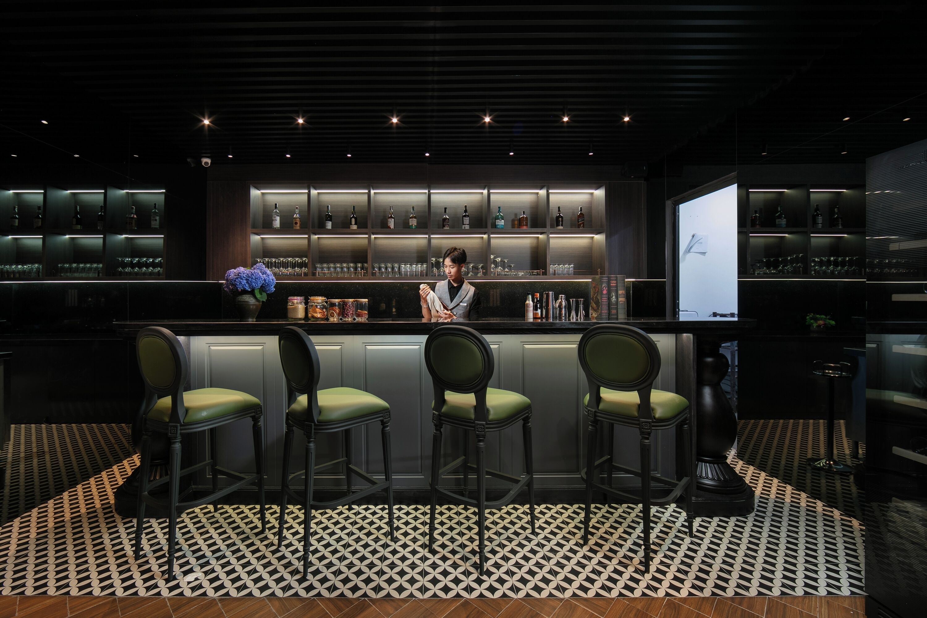 A bar area with a bartender, bar stools, shelves of bottles, and a patterned floor.