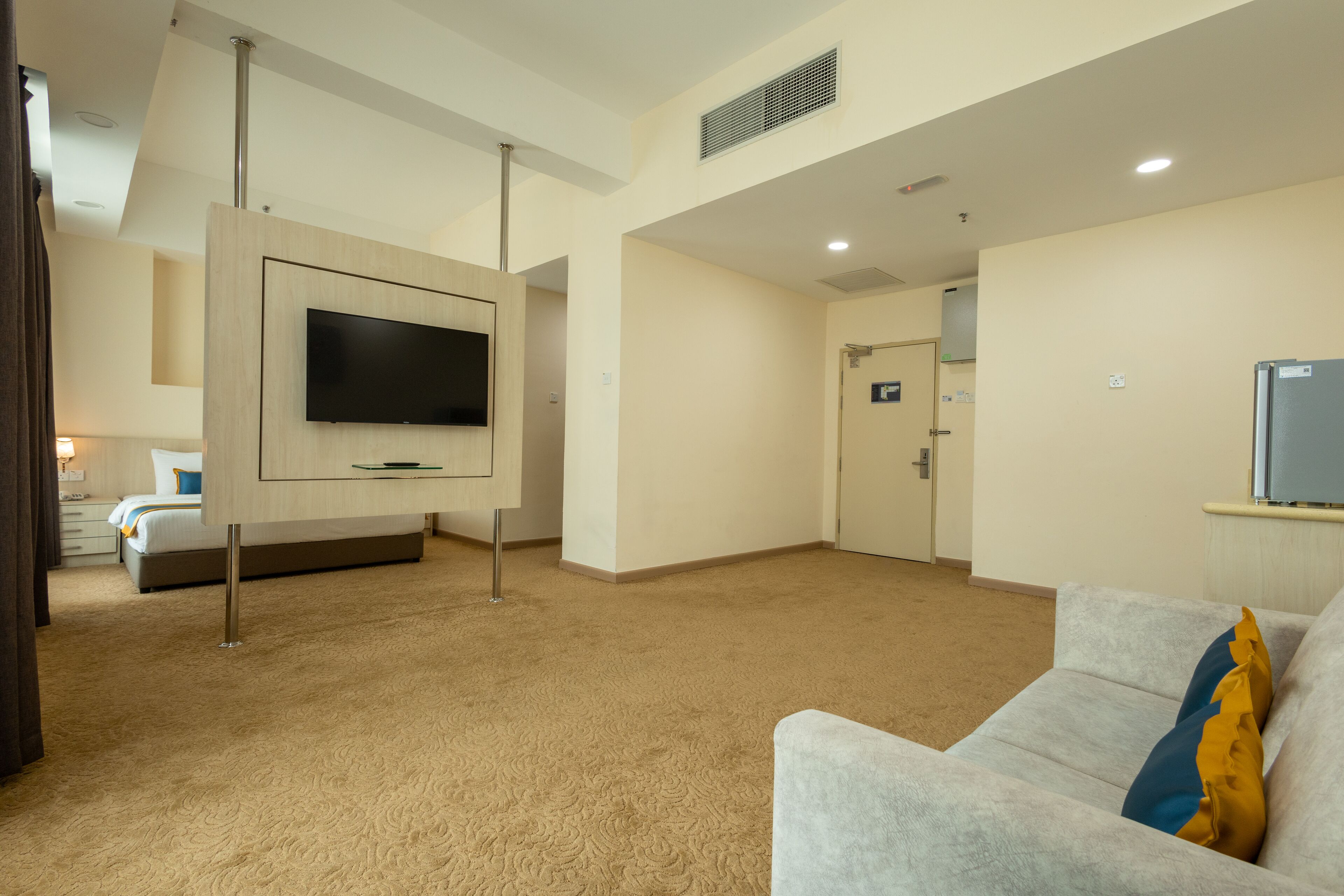 A modern hotel room with a large flat-screen TV mounted on a wooden panel, a grey sofa with a yellow pillow, and a beige carpet.