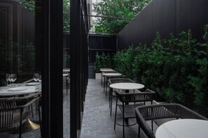 An outdoor dining area with wicker chairs and tables, surrounded by greenery and a dark wall. - Passa Hotel Bangkok (Bangkok)