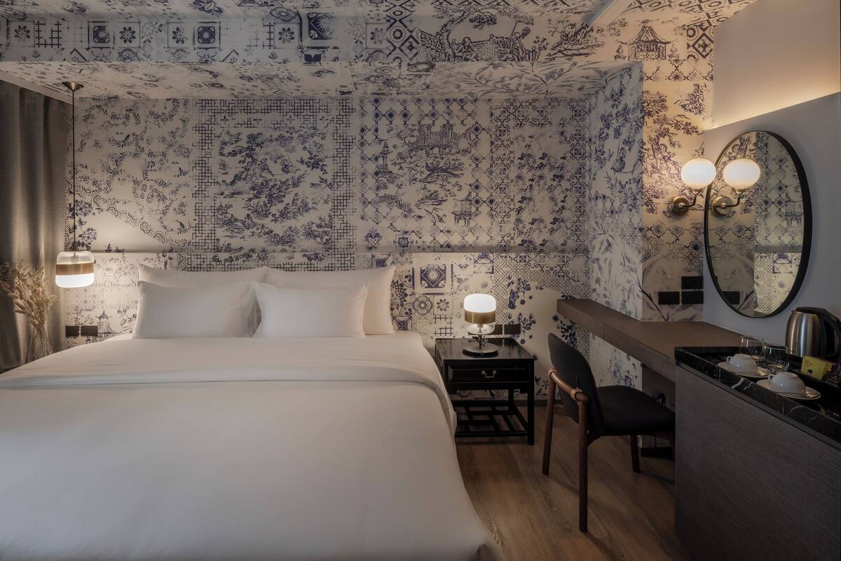 A hotel room with a large bed, a desk with a chair, a mirror, and a wall with patterned wallpaper.