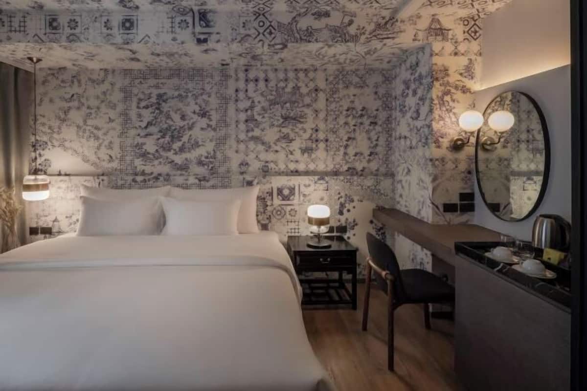 A hotel room with a large bed, bedside tables, a chair, a desk with a mirror, and a wall with patterned wallpaper.