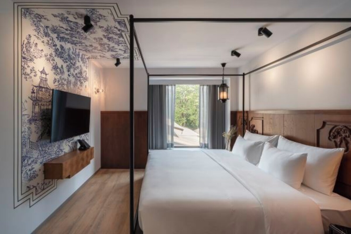 A modern hotel room with a large bed, a mounted TV, and a view of greenery outside.