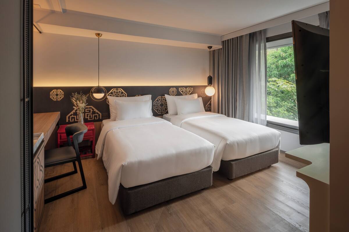 A hotel room with two beds, a desk, a chair, a TV, and a window with a view of greenery.