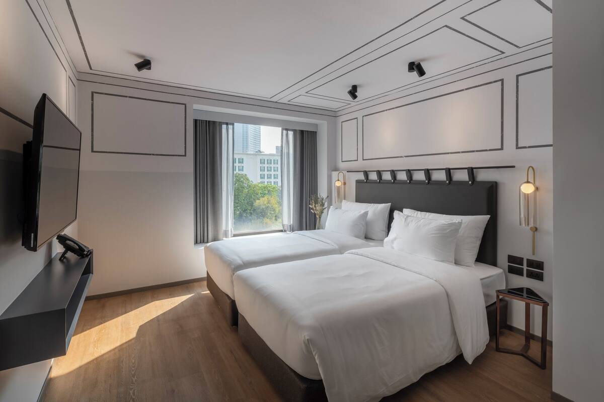 A modern hotel room with two beds, a flat-screen TV, and a large window offering a city view.