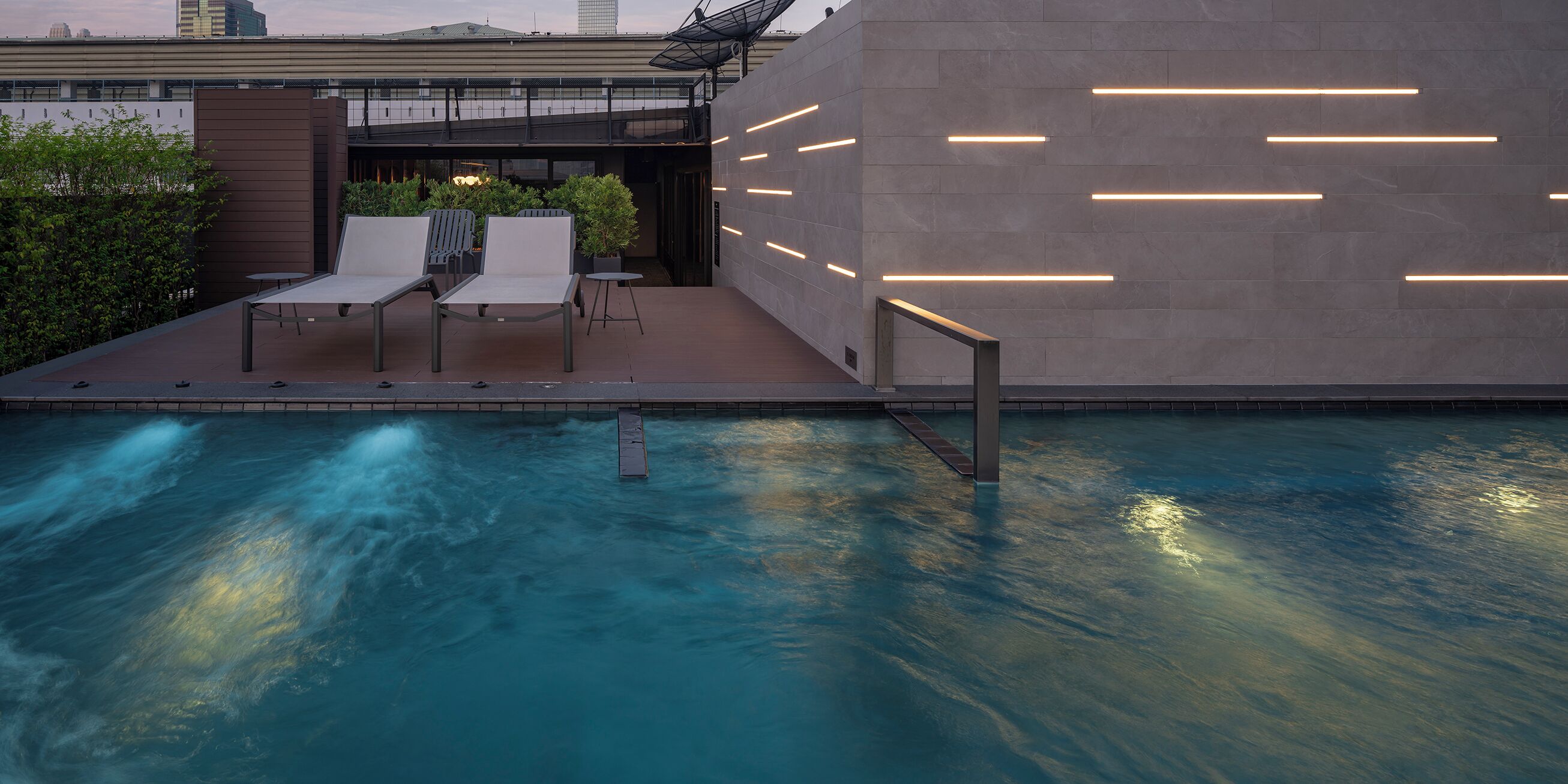 Ein moderner Poolbereich im Freien mit Liegestühlen und einem kleinen Tisch, umgeben von einer Steinwand mit beleuchteten Linien.