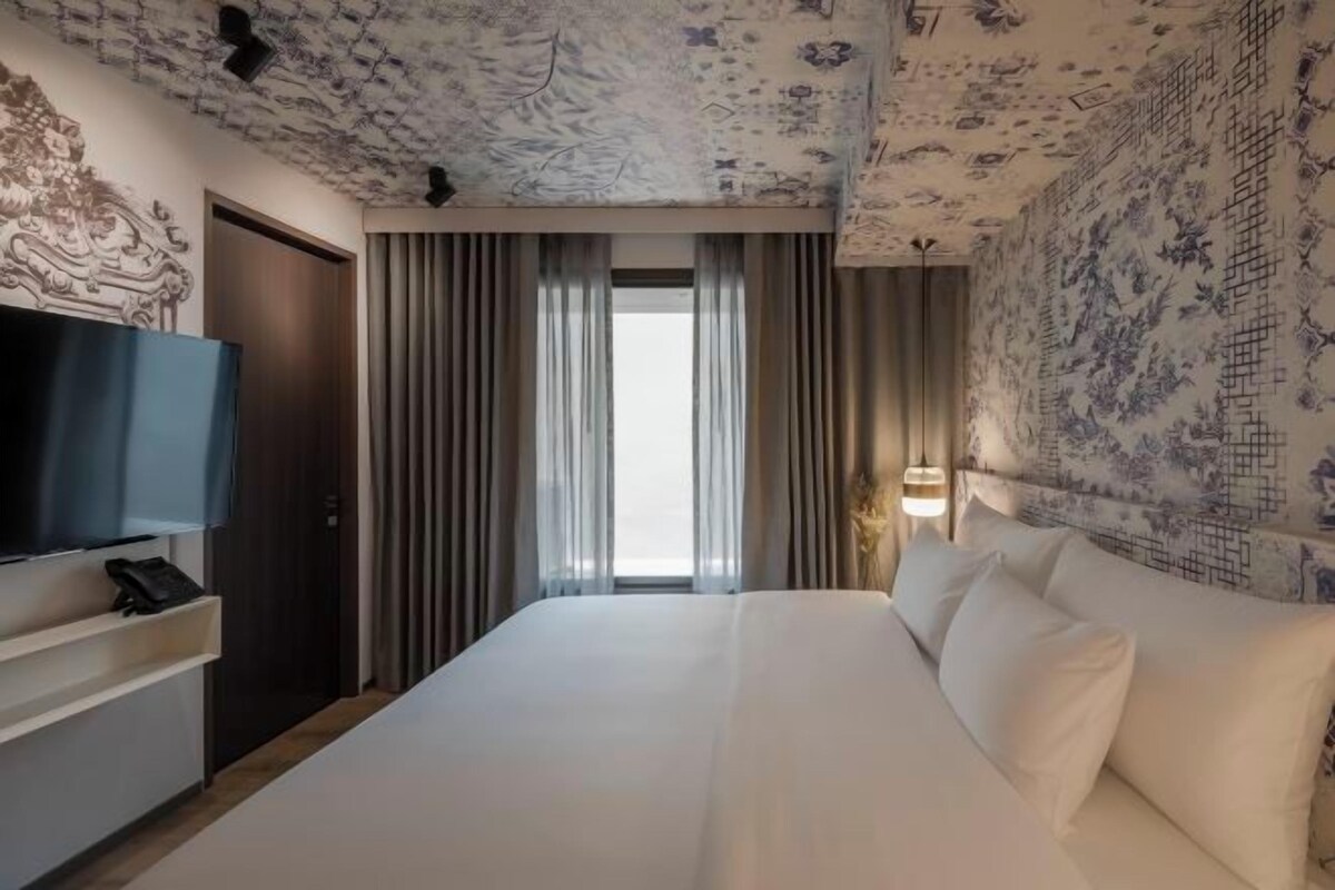 A hotel room with a large bed, a flat-screen TV, a desk with a phone, and a window with curtains.