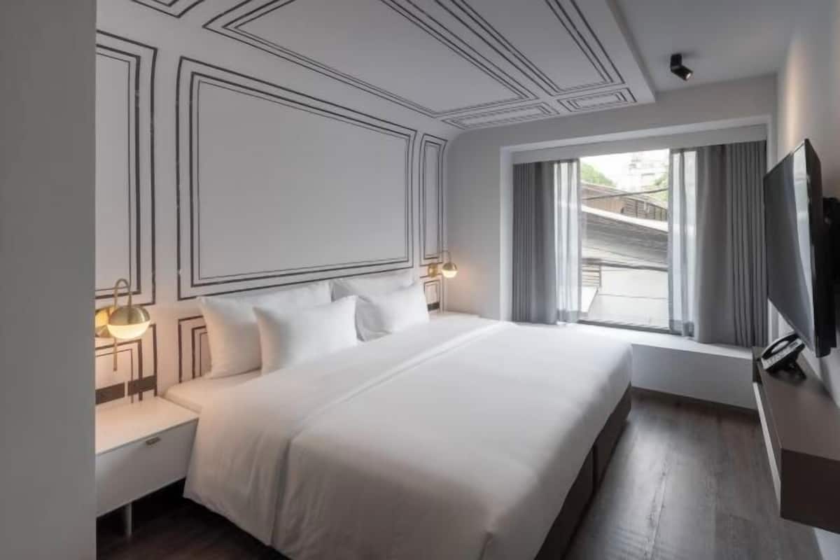 A hotel room with a large bed, a TV, a window with curtains, and a wall with geometric patterns.